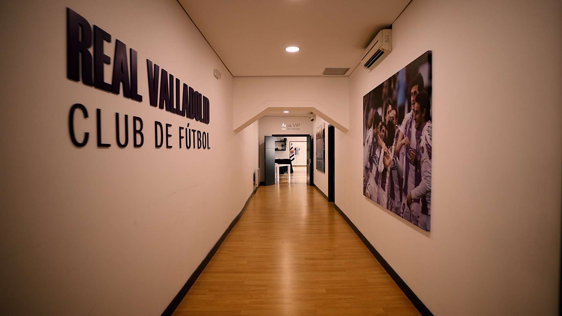 Pasillo con el suelo de madera, nombre del club en una pared y una foto del equipo en la otra, al fondo se ve una sala con mesas y sillas